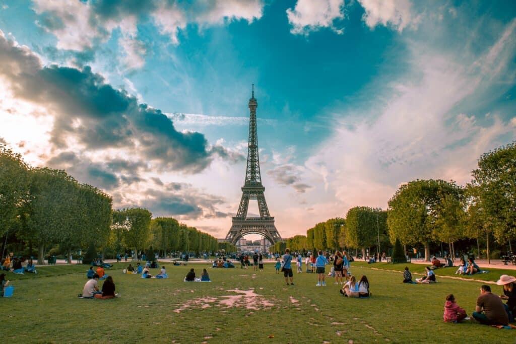 Campo verde em frente à Torre Eiffel com várias pessoas sentadas e passeando ao redor. Árvores ocupam as laterais do local.