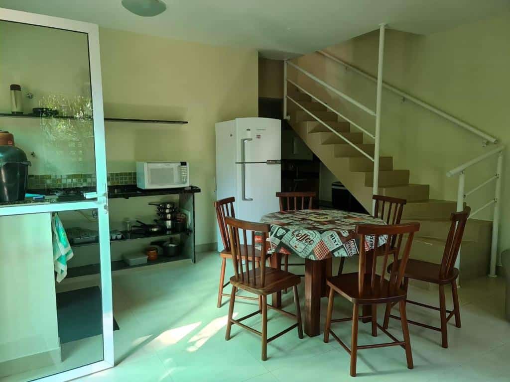 Cozinha de Itamambuca casas,suítes. Uma mesa com seis cadeiras no meio, no lado direito uma escada, no meio uma geladeira, do lado esquerdo uma pia com utensílios de cozinha e um micro-ondas.