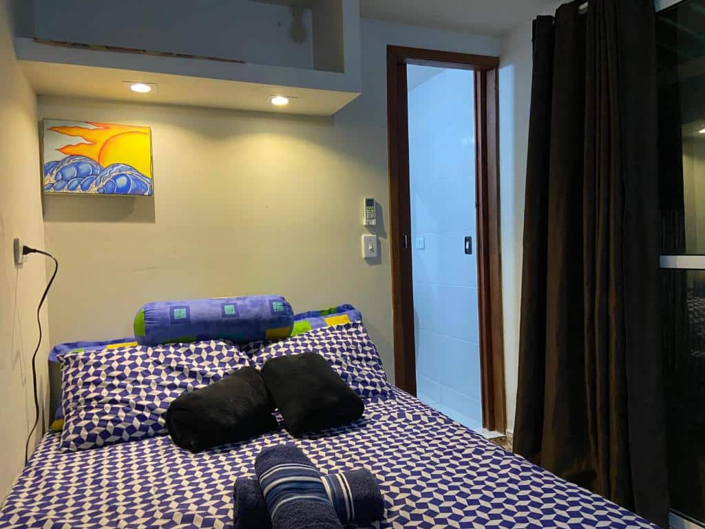 Quarto do JARDIM ITAMAMBUCA. Uma cama de casal do lado esquerdo, com várias almofadas e duas toalhas em cima, do lado direito uma cortina e a porta.