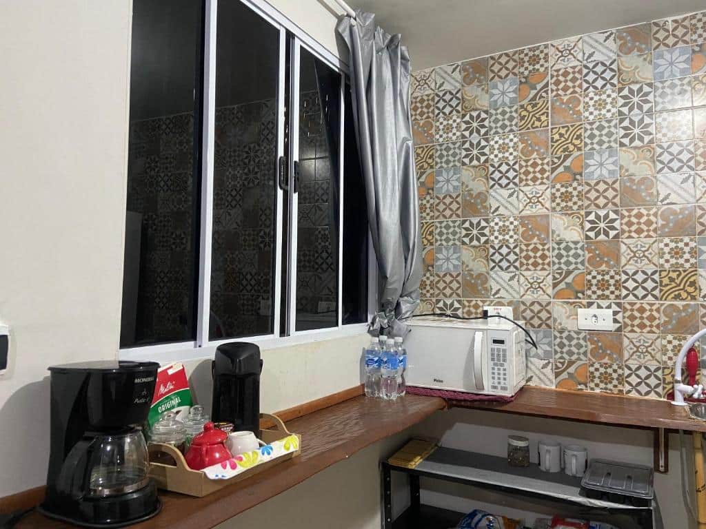 Cozinha do JARDIM ITAMAMBUCA. Um balcão com cafeteira, coisas de café, no fundo um micro-ondas, garrafa de água, em cima uma janela e em baixo uma estante.