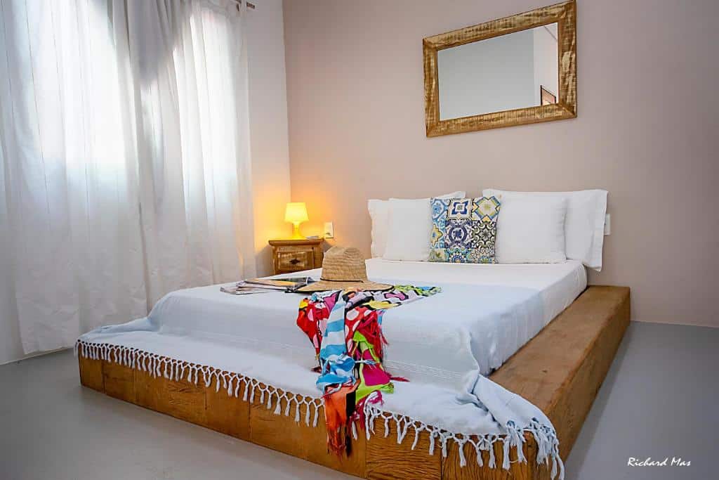Quarto do Jardim Secreto. Uma cama de casal no meio, do lado esquerdo uma cômoda com abajur, uma janela com cortinas fechadas. Em cima da cama um espelho. Foto para ilustrar post sobre airbnb em Morro de São Paulo.