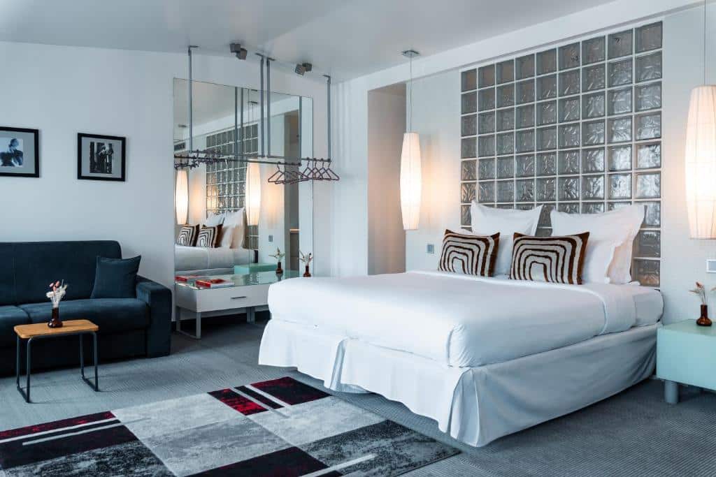 Quarto do Kube Hotel Paris - Ice Bar, um dos hotéis com estacionamento gratuito em Paris. Uma cama de casal está encostada na parede direita com mesinhas de cabeceira e abajures dos dois lados. Na parede esquerda ao lado da cama há um cabideiro em frente de uma parede espelhada e acima de um móvel branco. Ao lado do espelho fica um sofá e uma mesinha. Um tapete estampado está em frente à cama.