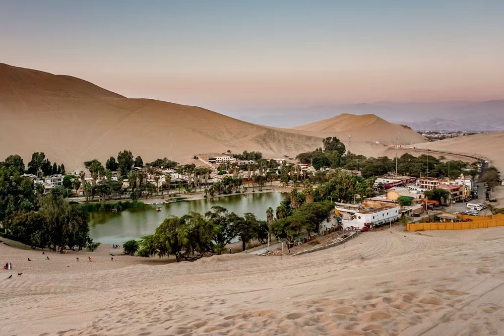 Lago rodeado de árvores e casas, com um deserto de areia a sua volta. O local é o Lago de Huacachina, no Peru. Foto: Civitatis
