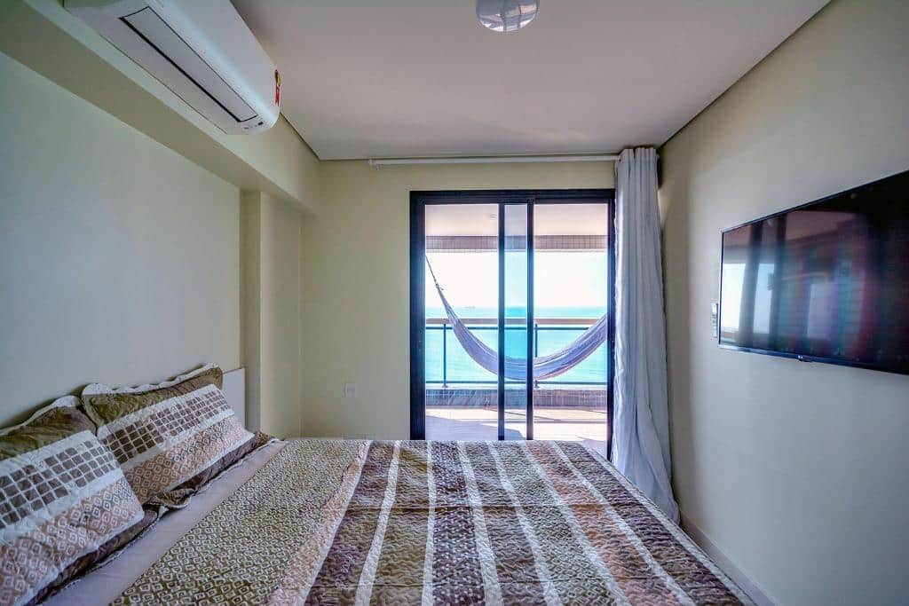 Quarto do airbnb Landscape Beira Mar Platinum. Uma cama de casal está no canto e de frente para uma tv. Ao fundo está a porta para a varanda, que conta com uma rede e vista para o mar.
