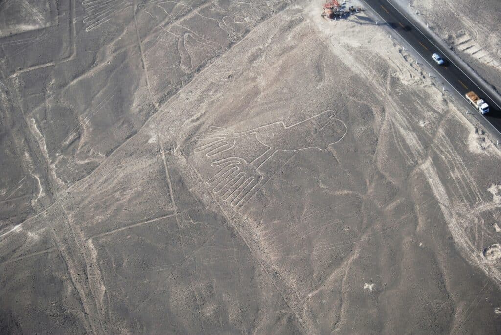 Vista aérea das linhas de Nazca, com uma estrada com um carro e um caminhão andando, à direita. Na imagem, há um campo extenso de areia com desenhos históricos.Representa o que fazer no Peru