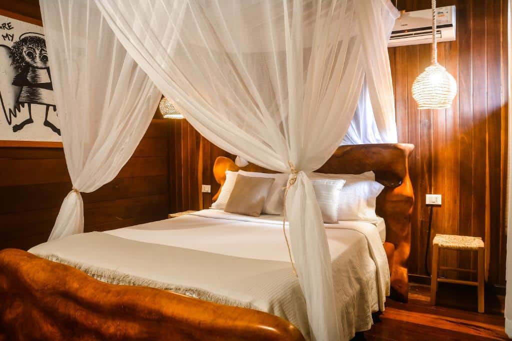 Quarto do Local Brasil House. Uma cama de casal no meio, de cada aldo bancos de madeira e palha e luminárias. Em cima um ar-condicionado e véu na cama.