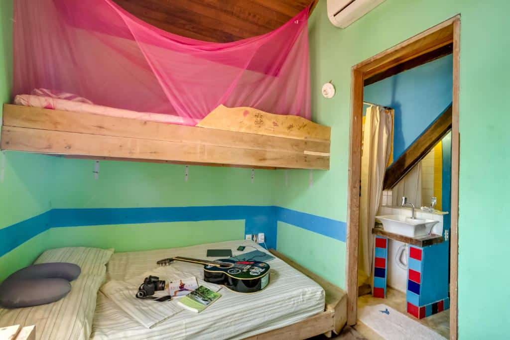 Há uma cama beliche à esquerda, no Mais que Nada Itacaré Hostel.  Na cama de baixo há um violão, livros e câmera fotográfica em cima. À direita vemos uma porta para o banheiro e a pia dele.