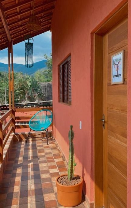 Frente do quarto do Mandacaru Flats Itamambuca. A porta do lado direito com um cacto de decoração, no fundo a janela, em baixo uma cadeira e a ponta da varanda.