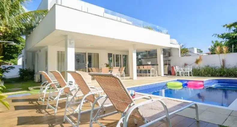 Vista de espreguiçadeiras encarando a piscina da Mansão em Barequeçaba. Ao fundo está a casa branca e uma mesa posta com cadeiras. Há boias na piscina desse airbnb em Barequeçaba.