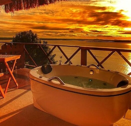 Foto da varanda do Mar e Morro Flats para ilustrar post sobre airbnb em Morro de São Paulo. Uma banheira de hidromassagem do lado direito, do lado esquerdo uma mesa de madeira. Vista para o mar e o pôr do sol.