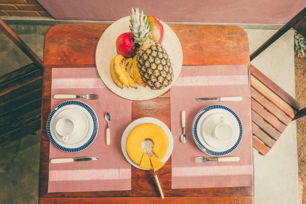 Mesa de café da Vila Prosperidade Trancoso com duas xicáras com dois pratos em cada lado com talheres no centro um prato com bolo e do outro lado um prato com frutas, maçã, mamão, banana e abacaxi.