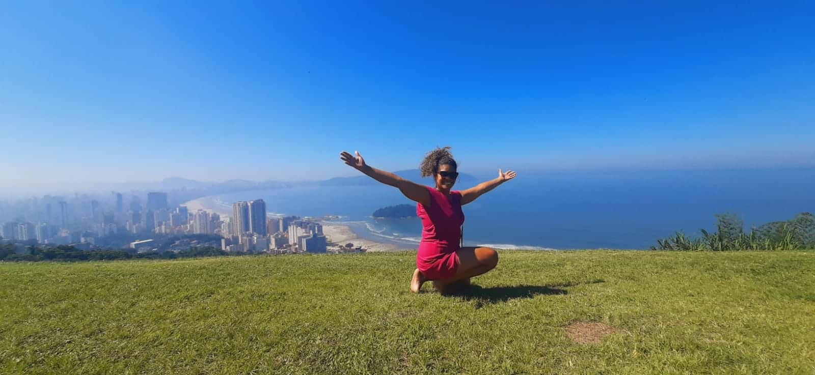 Vista do Morro Voturuá. Na frente uma mulher agachada com os braços abertos, atrás a vista do morro. Do lado direito o mar, do lado esquerdo os prédios.