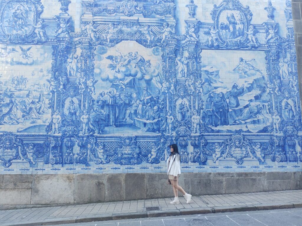 Mulher andando durante o dia e ao fundo o mural de azulejos da Igreja. 