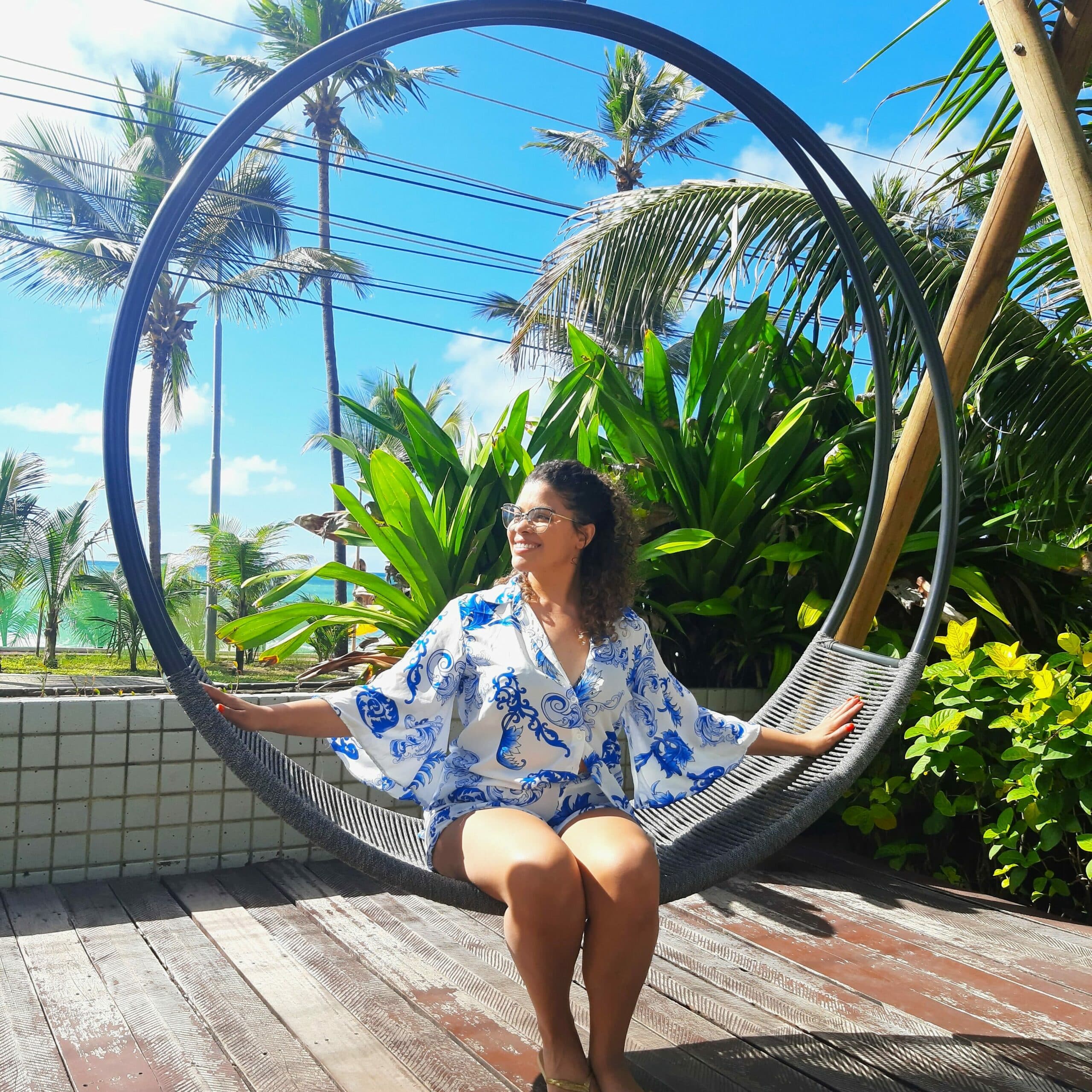 Mulher sentada em um balanço redondo do Ritz Lagoa da Anta com os braços abertos e apoiados no balanço. Há folhas verdes à direta e vista do mar ao fundo com coqueiros balançando