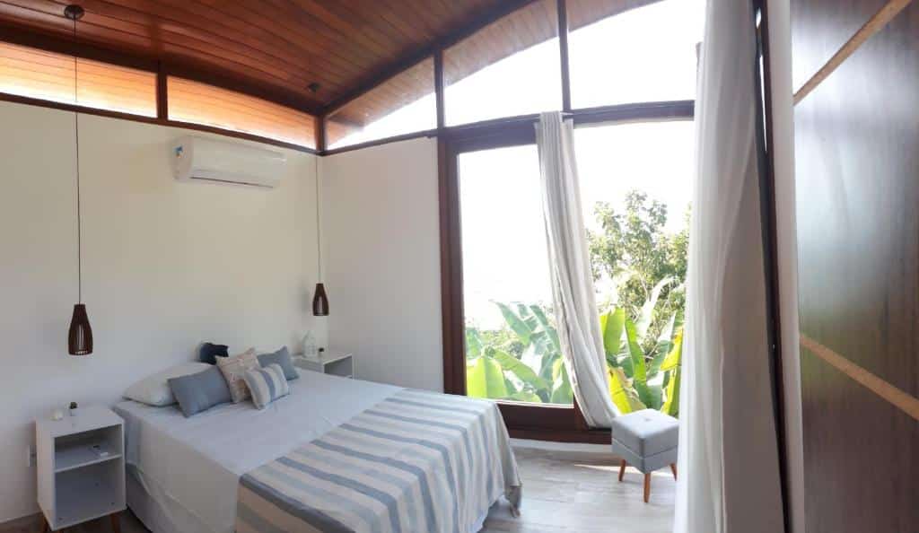Quarto da Novissima Casa. Do lado esquerdo uma cama de casal com uma cômoda de cada lado e abajur. Em cima um ar-condicionado. No fundo uma parede de vidro. Foto para ilustrar post sobre airbnb em Morro de São Paulo.