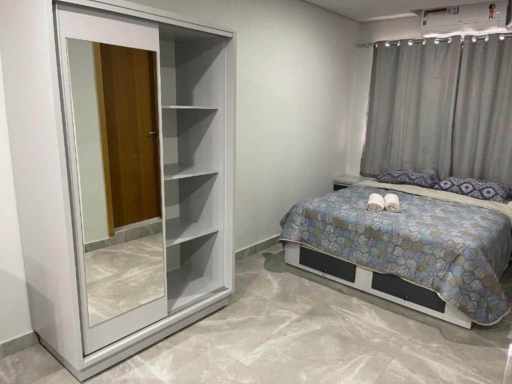 Quarto do Novo apartamento Studio. Um guarda-roupa com porta de correr e um espelho está à esquerda, e à direita há uma cama de casal, com uma janela e ar-condicionado atrás. Está ilustrando o post sobre airbnb em Foz do Iguaçu.