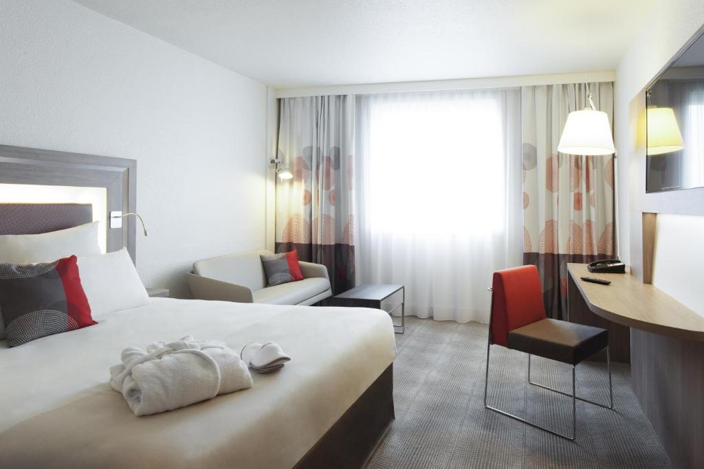 Quarto do Novotel Paris 17, um dos hotéis Novotel em Paris. Uma cama de casal está encostada na parede do lado esquerdo e conta com mesinhas de cabeceira e luminárias dos dois lados, assim como chinelos e um roupão aos pés. Logo em frente fica uma mesa de trabalho com cadeira, abajur, telefone e uma TV na parede acima. Ao lado da cama há um sofá com mesinha, e ao lado fica uma janela com cortinas.