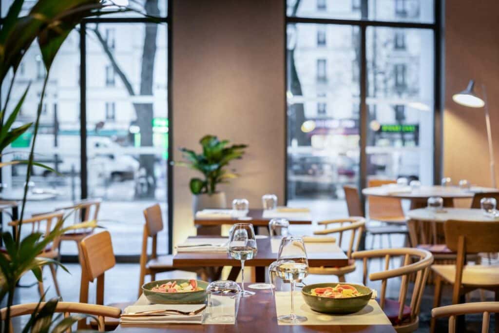 Restaurante do Novotel Paris 20 Belleville, onde várias mesas com cadeiras estão dispostas. Há janelas na parede ao fundo, e alguns vasos de plantas estão dispostos pelo local.