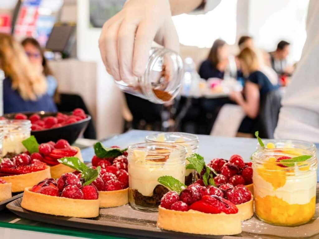 Close em tortinhas de framboesa e potes de doces numa mesa do Novotel Paris Centre Bercy. Uma mão despeja algo em um pote, e ao fundo é possível ver pessoas almoçando.