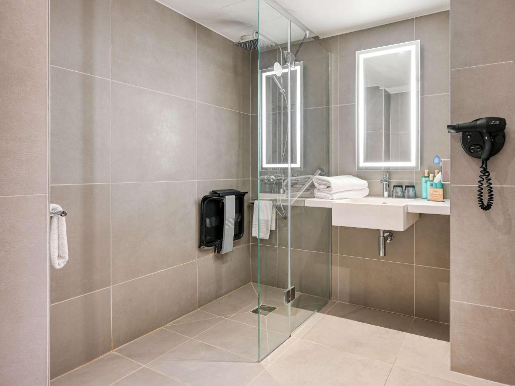 Banheiro adaptado para PCDs no Novotel Paris Centre Bercy. Um amplo box de vidro tem barras de apoio e assento de banho do lado de dentro. Ao lado fica uma pia com amenidades de banho, secador de cabelo, toalhas e espelho.