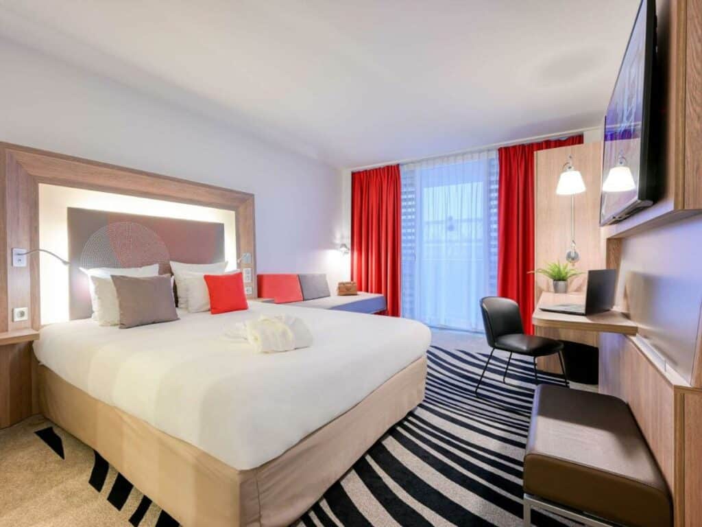 Quarto do Novotel Paris Centre Bercy, um dos hotéis Novotel em Paris. Uma cama de casal está encostada na parede do lado esquerdo e conta com mesinhas de cabeceira e luminárias dos dois lados, assim como um roupão dobrado aos pés. Logo em frente fica uma TV com um banco abaixo. Ao fundo há um sofá na mesma parede da cama que encara uma mesa de trabalho com cadeira, luminária, notebook e vaso de plantas na parede oposta. Há uma janela com cortinas ao lado do sofá.