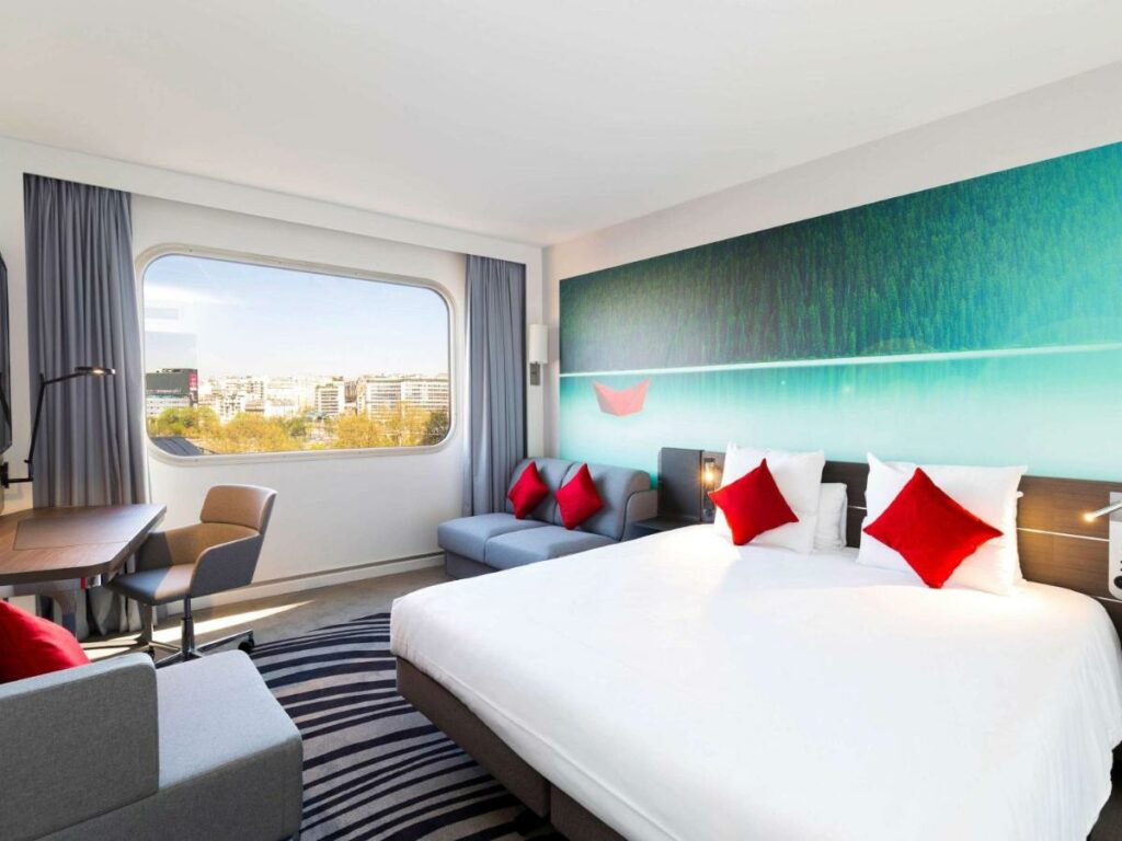 Quarto do Novotel Paris Centre Tour Eiffel, um dos hotéis Novotel em Paris. Uma cama de casal está encostada na parede do lado direito e conta com mesinhas de cabeceira e luminárias dos dois lados. Ao lado esquerdo há um sofá-cama e uma mesa de trabalho com cadeira e luminária. Ao lado há uma janela com vista para a cidade e cortinas. Logo em frente à cama há uma TV na parede e um sofá abaixo.