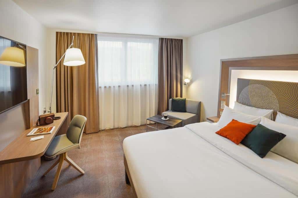 Quarto do Novotel Paris Gare De Lyon, um dos hotéis Novotel em Paris. Uma cama de casal está encostada na parede do lado direito e conta com mesinhas de cabeceira e luminárias dos dois lados. Logo em frente fica uma mesa de trabalho com cadeira, abajur e TV na parede acima. Ao fundo há uma área de estar com mesinha e poltrona, além de uma janela com cortinas.