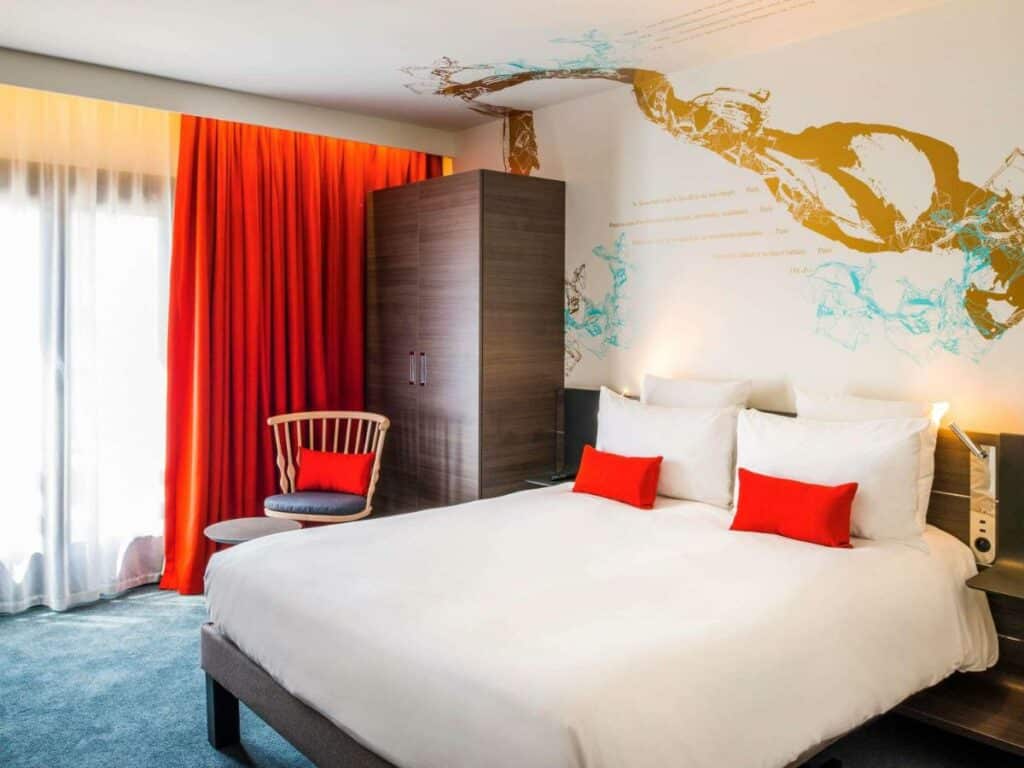 Quarto do Novotel Paris Les Halles, um dos hotéis 4 estrelas em Paris. Uma cama de casal está encostada na parede do lado direito e conta com mesinhas de cabeceira e luminárias dos dois lados. Ao lado esquerdo há uma cadeira em frente à janela com cortinas. Um guarda-roupas fica logo atrás da cadeira.