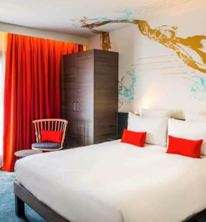 Quarto do Novotel Paris Les Halles, um dos hotéis Novotel em Paris. Uma cama de casal está encostada na parede do lado direito e conta com mesinhas de cabeceira e luminárias dos dois lados. Ao lado esquerdo há uma cadeira em frente à janela com cortinas. Um guarda-roupas fica logo atrás da cadeira.