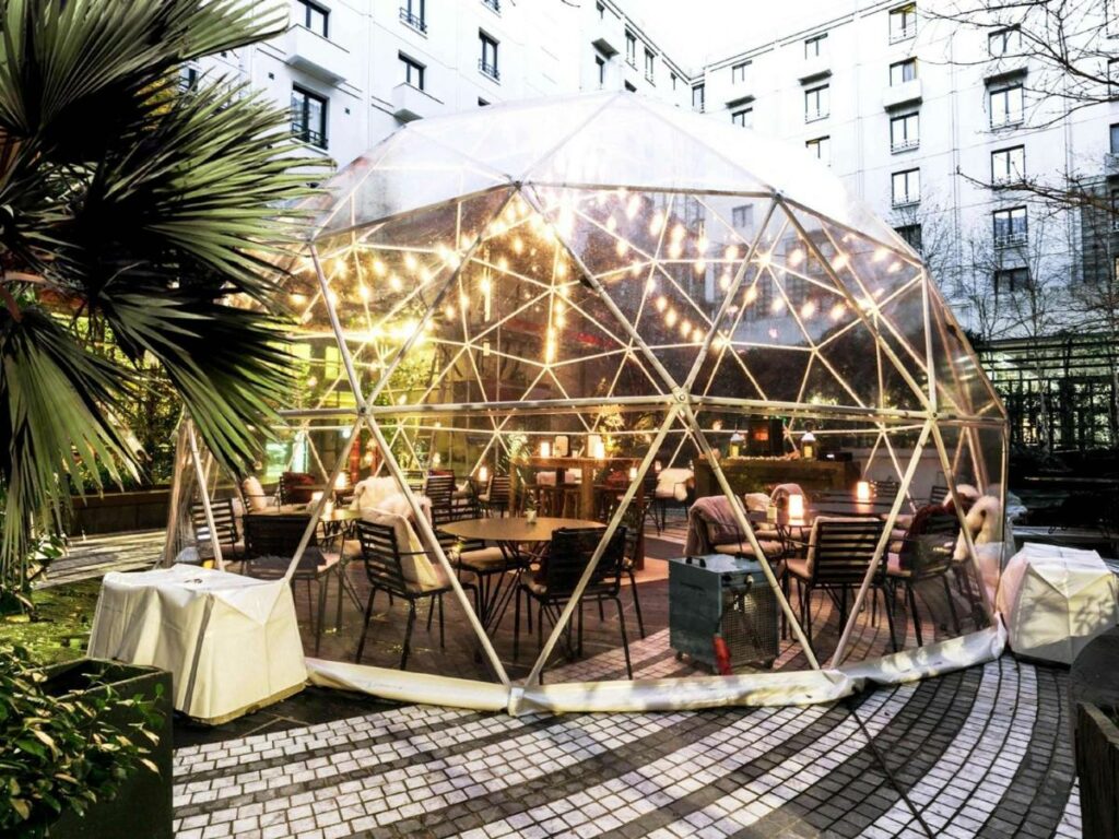 Restaurante do Novotel Paris Les Halles localizado dentro de uma bolha de vidro com iluminações delicadas. Ali dentro há algumas mesas e cadeiras, e do lado de fora ficam plantas.