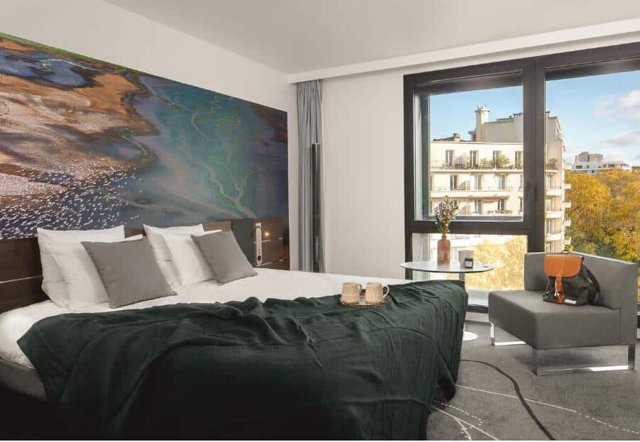 Quarto do Novotel Paris Porte Versailles, um dos hotéis Novotel em Paris. Uma cama de casal está encostada na parede do lado esquerdo e tem mesinhas de cabeceira e luminárias dos dois lados. Ao lado direito da imagem fica uma área de estar com mesinha e poltrona logo em frente à uma ampla janela com vista para a cidade.