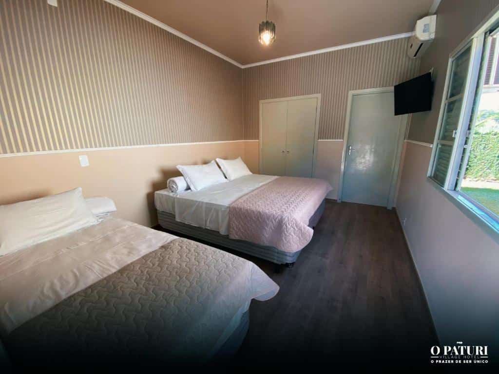 Quarto espaçoso no O Paturi – Village Hotel. Há duas camas de casal, uma do lado da outra, um guarda-roupa na parede do fundo e uma janela e TV na frente delas. É uma indicação de hotéis perto da Dutra.