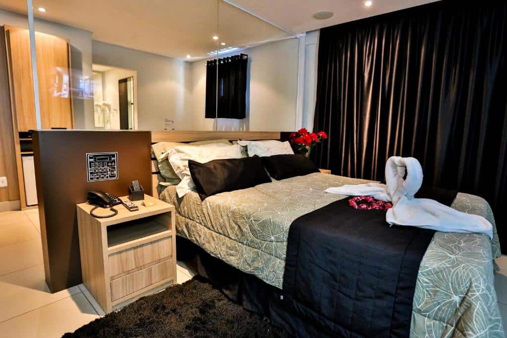 Quarto do Palais Hotel. Uma cama de casal no meio, com uma decoração de coração com toalhas. De cada lado uma cômoda com telefone e um vaso de flor. Um vidro separando a outra parte do quarto. Foto para ilustrar post sobre pousadas em São Vicente.