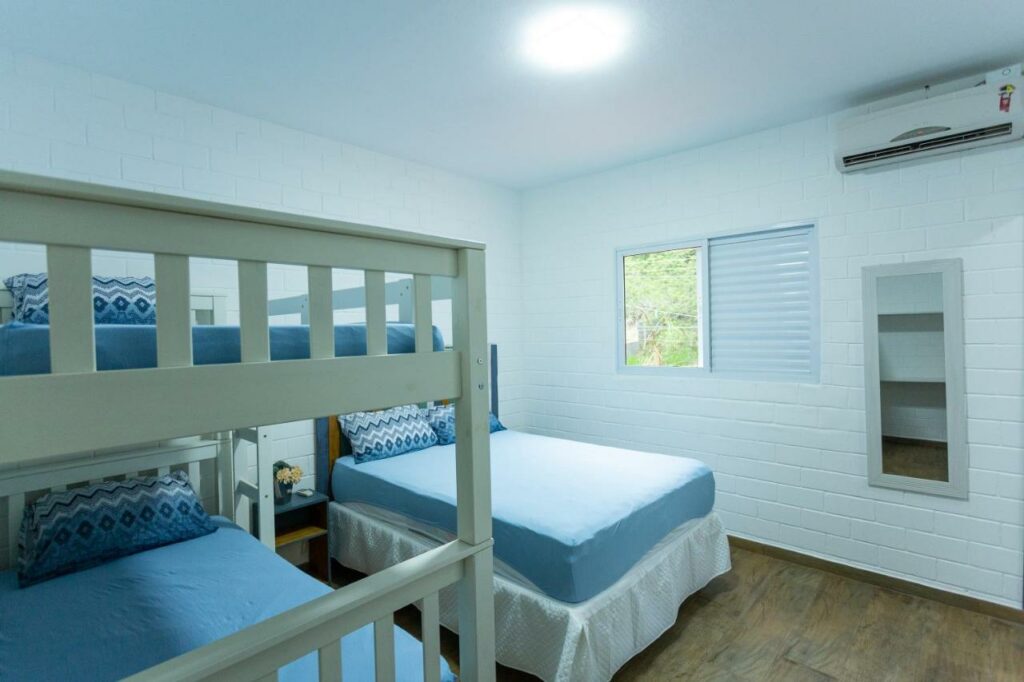 Quarto do Paraíso Azul, um dos airbnb em Barequeçaba. Uma beliche está encostada na parede ao lado esquerdo e tem uma cama de casal ao lado. Há mesinhas de cabeceira dos dois lados da cama, e uma janela na parede ao lado. Na mesma parede da janela há um espelho e um aparelho de ar-condicionado.