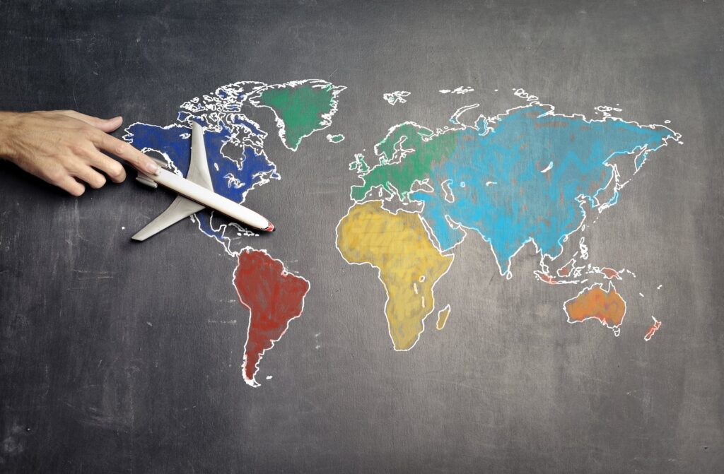 Um quadro negro com o mapa mundi desenhado com giz colorido, e no topo do lado esquerdo há uma mão segurando um avião de brinquedo