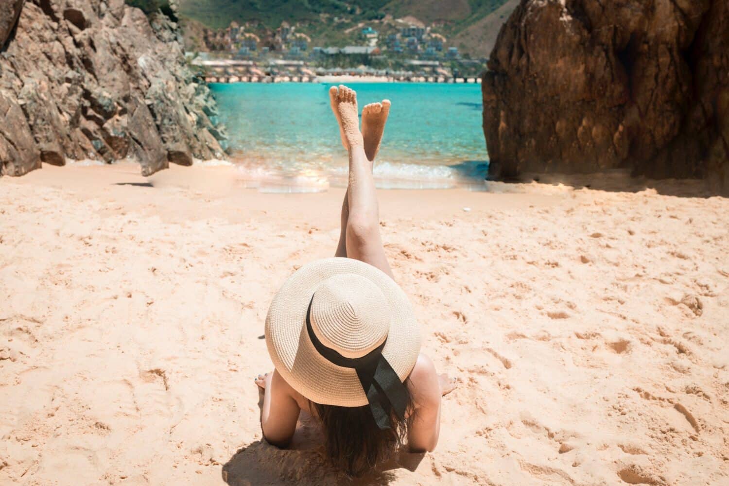 Uma mulher deitada em uma areia clara com um chapéu na cabeça olhando para um mar muito azul, para representar o melhor seguro viagem