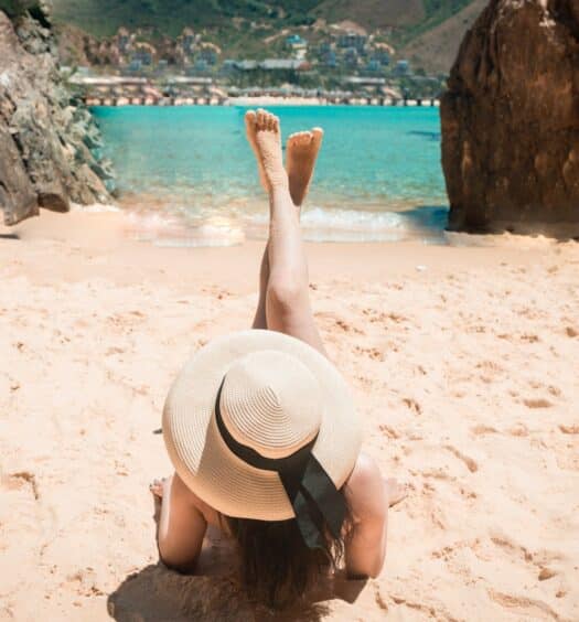 Uma mulher deitada em uma areia clara com um chapéu na cabeça olhando para um mar muito azul, para representar o melhor seguro viagem