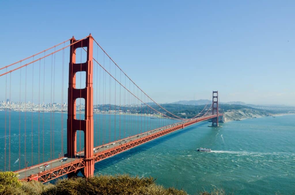 A Ponte Golden Gate em São Francisco, a estrutura é vermelha e muito alta, corta um amplo rio com uma embarcação passando