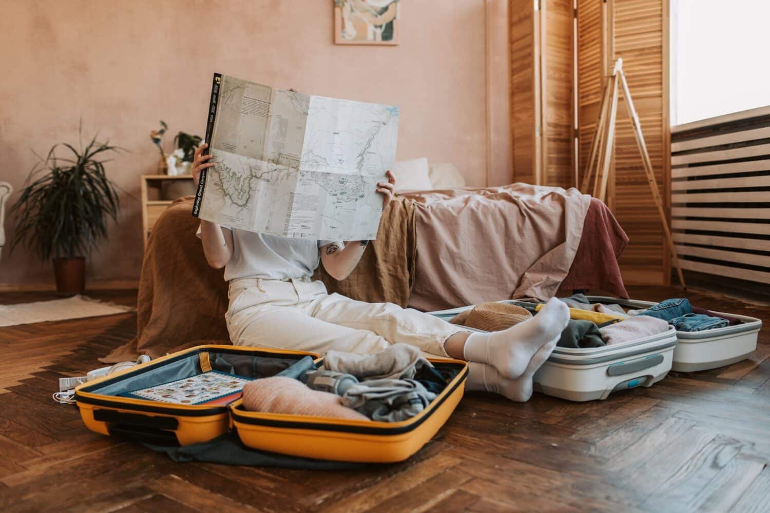 Uma mulher sentada no chão do quarto segurando um mapa em mãos, ao redor dela há duas malas abertas com itens dentro, para representar o seguro viagem para quem viaja muito