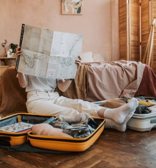 Uma mulher sentada no chão do quarto segurando um mapa em mãos, ao redor dela há duas malas abertas com itens dentro, para representar o seguro viagem para quem viaja muito