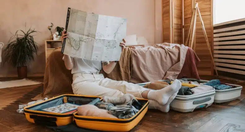 Uma mulher sentada no chão do quarto segurando um mapa em mãos, ao redor dela há duas malas abertas com itens dentro, para representar o seguro viagem para quem viaja muito