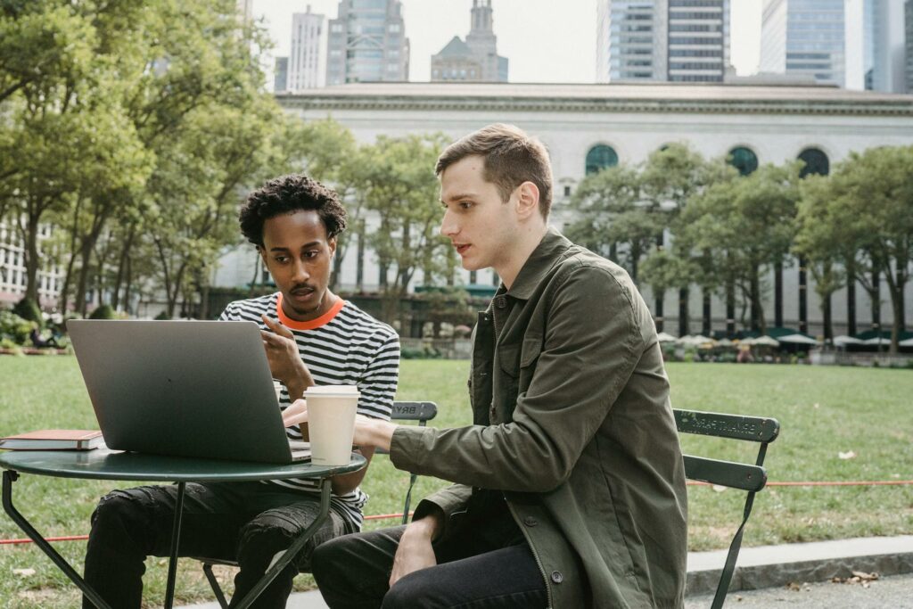 Dois rapazes, um negro de camiseta listrada e outro branco com uma jaqueta verde, estão sentados em um local aberto com árvores, em frente deles há uma mesa com um notebook aberto e ambos estão olhando para o eletrônico