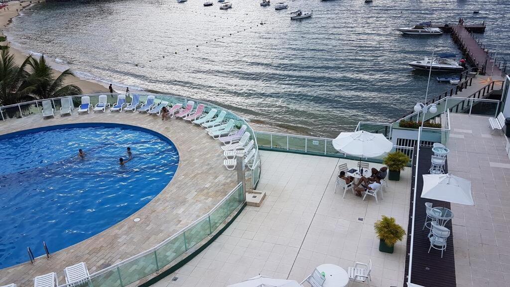 Área externa do airbnb Angra inn. No canto esquerdo há uma piscina redonda com espreguiçadeiras em volta, no lado direito há mesas com cadeiras e guarda-sóis e ao fundo da imagem está o mar.