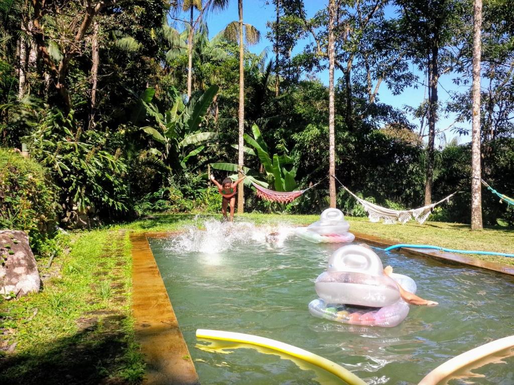 Piscina do airbnb Chalé Gaia. A piscina é retangular e está centralizada na imagem. Em volta da piscina há uma grande área verde e já dentro da piscina há boias e espaguetes. Há também uma criança pronta para pular na piscina. Imagem para ilustrar o post airbnb em Itatiaia.