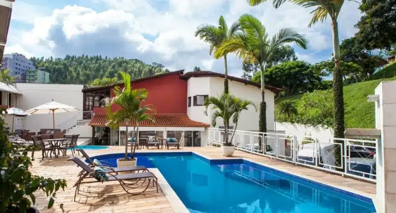 Área externa do Mundial Parque Hotel. Uma piscina com formato em L está na frente da imagem e atrás há uma construção e uma área verde, para representar hotéis em Viçosa.