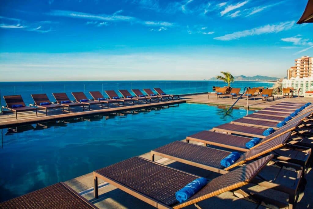 Vista da piscina do Windsor Oceanico Hotel durante o dia com cadeiras a frente no meio a piscina e ao fundo o mar. Representa airbnb na Barra da Tijuca.