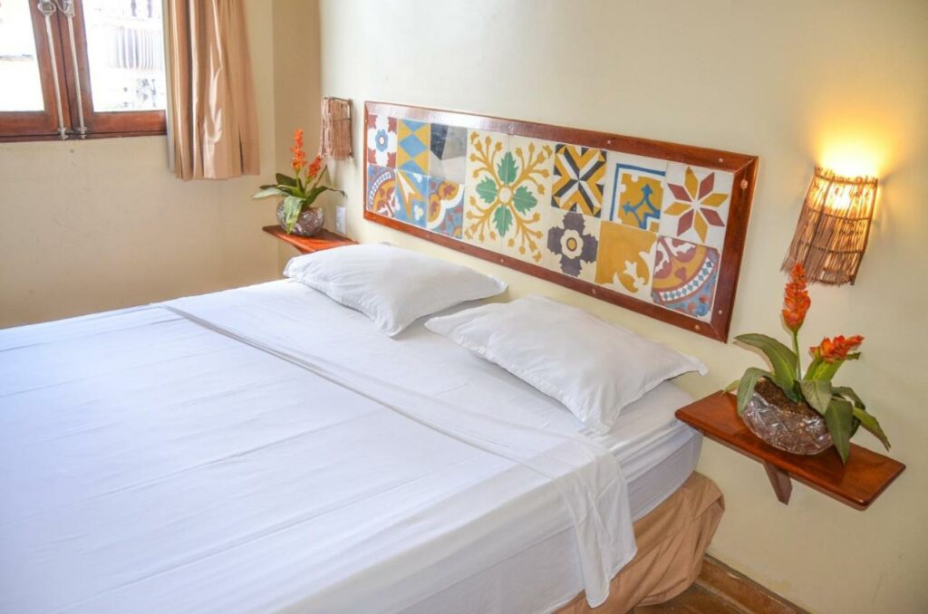 Quarto da Pousada Fortal Villa Praia. Uma cama de casal está no meio e existem duas prateleiras com vasos de flor em cada lado.
