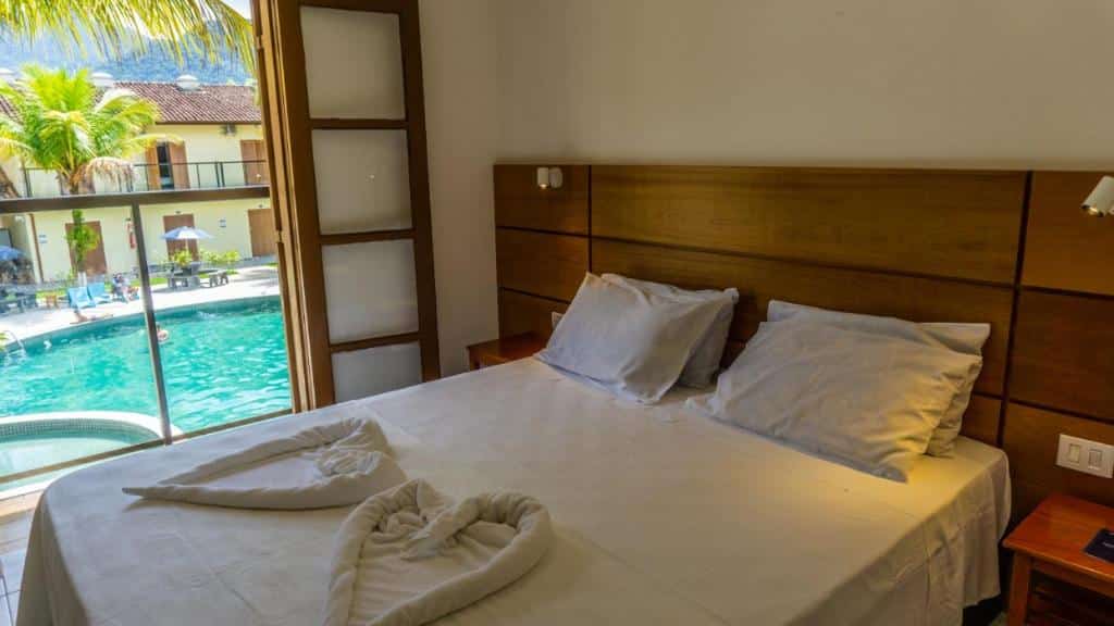 Cama de casal em quarto da Pousada Ilha Vitoria, com toalhas em formato de coração, e vista da piscina pela varanda