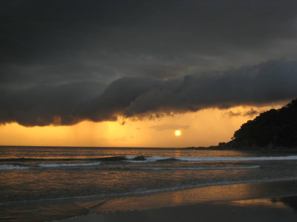 Vista da Praia da Barra do Sahy durante o final do dia com o mar a frente e ao fundo o sol de com várias nuvens em volta.