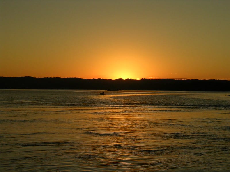 O sol está se pondo na Praia da Coroa, em Itacaré. O céu está tomado pela cor laranja, que reflete nas águas. Lá no fundo, há um pequeno barco.